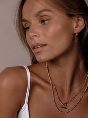 A woman with gold hoop earrings and layered gold necklaces, wearing a white top, looks to the side, accessorized with a Crossing Twisted Chain.