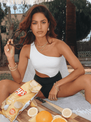 A woman in a swimsuit sits with a bag of chips, lemons, oranges, a knife, and a Chunky Link and Round Hook Toggle on a wooden board.